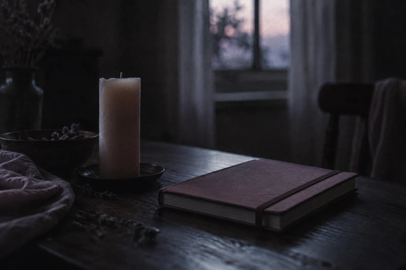 An unlit wax candle and a closed notebook on a dark table, one faint rim of window light